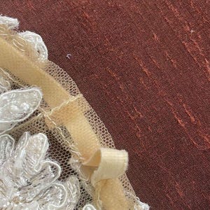 May include: Close-up of a cream-colored lace applique with beaded accents. A pale yellow ribbon is attached to the lace, forming a small bow. The background is a textured, reddish-brown fabric.