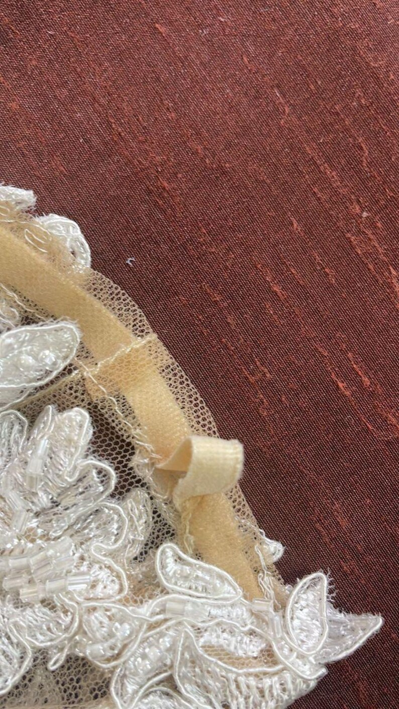 May include: Close-up of a cream-colored lace applique with beaded accents. A pale yellow ribbon is attached to the lace, forming a small bow. The background is a textured, reddish-brown fabric.