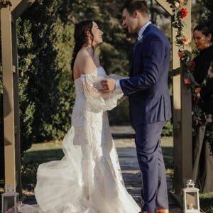 May include: A wedding ceremony scene featuring a bride in a white, off-the-shoulder lace gown and a groom in a navy blue suit. They stand under a wooden arch decorated with flowers. The setting is outdoors, with greenery in the background.