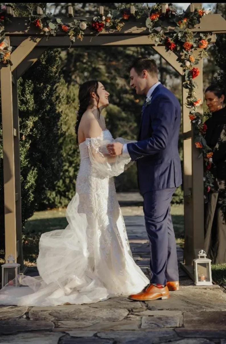 May include: A wedding ceremony scene featuring a bride in a white, off-the-shoulder lace gown and a groom in a navy blue suit. They stand under a wooden arch decorated with flowers. The setting is outdoors, with greenery in the background.