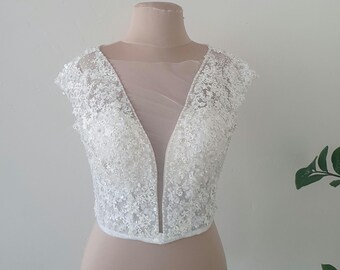 May include: A white sleeveless bridal bodice featuring intricate floral lace embroidery and beadwork. The garment has a deep V-neckline with a sheer illusion mesh panel extending to the shoulders and cap sleeves, creating a structured silhouette.
