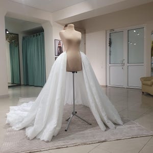 May include: A white bridal skirt with intricate lace detailing is draped over a mannequin. The skirt is displayed on a silver stand in a well-lit room with a neutral-toned rug. The skirt's train extends across the floor.