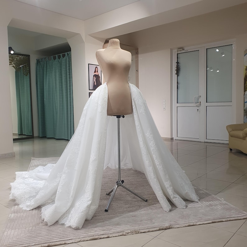 May include: A white bridal skirt with intricate lace detailing is draped over a mannequin. The skirt is displayed on a silver stand in a well-lit room with a neutral-toned rug. The skirt's train extends across the floor.