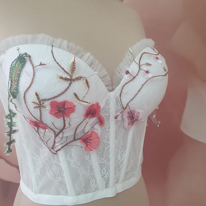 May include: White lace corset with floral embroidery in shades of pink, green, and brown. The corset has a sweetheart neckline and a boned bodice.