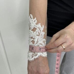 May include: White lace bridal sleeve with floral design. A pink measuring tape shows the sleeve's circumference is approximately 2 inches. The sleeve is made of sheer fabric.