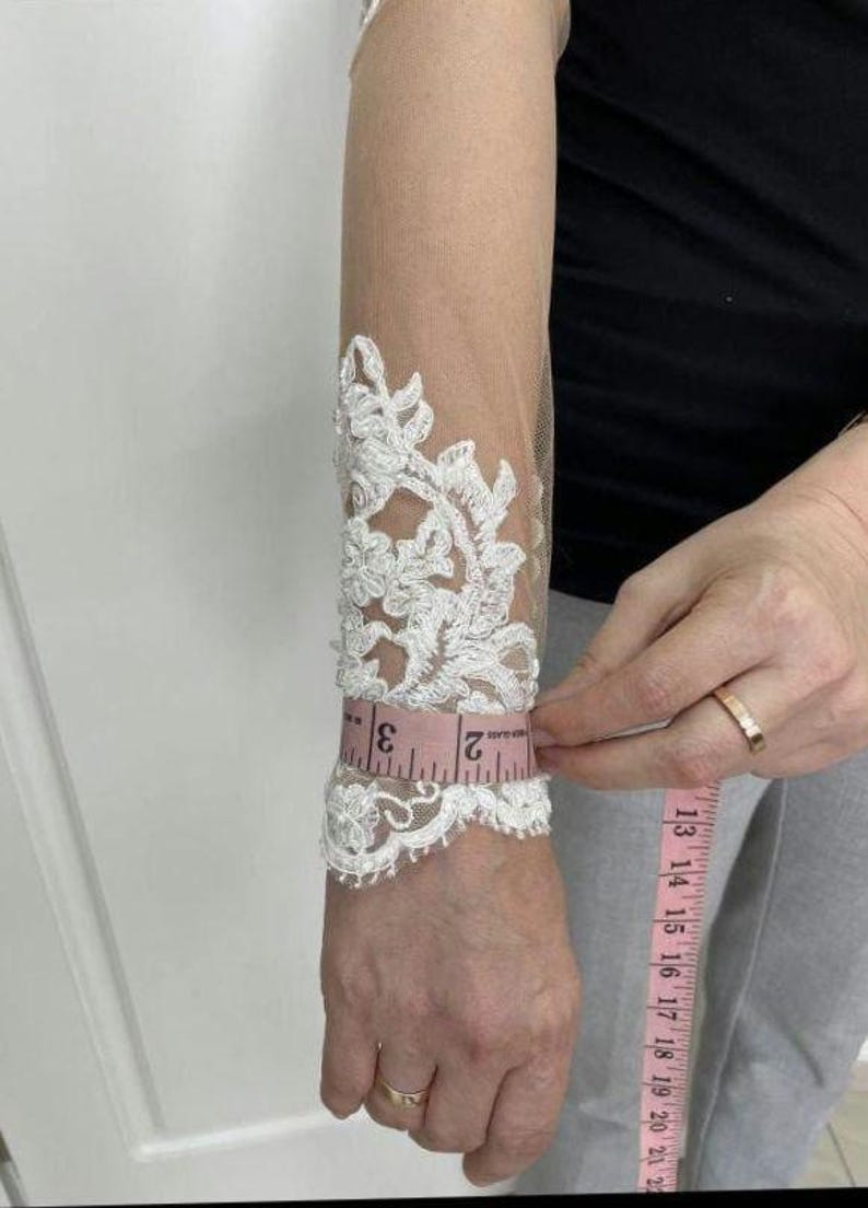 May include: White lace bridal sleeve with floral design. A pink measuring tape shows the sleeve's circumference is approximately 2 inches. The sleeve is made of sheer fabric.