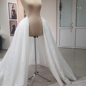 May include: A white bridal skirt with lace detailing is draped over a mannequin. The skirt is displayed on a metal stand in a well-lit room. Two framed pictures hang on the wall behind the mannequin.