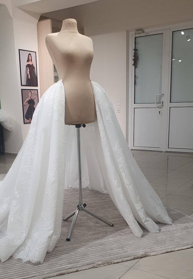 May include: A white bridal skirt with lace detailing is draped over a mannequin. The skirt is displayed on a metal stand in a well-lit room. Two framed pictures hang on the wall behind the mannequin.