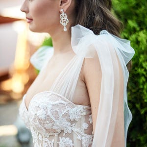 Könnte beinhalten: Weißes Hochzeitskleid mit floralem Spitzenmieder und Tüll-Overlay. Das Kleid hat einen herzförmigen Ausschnitt und zarte Träger. Die Braut trägt ein funkelndes Haarteil und Ohrringe.