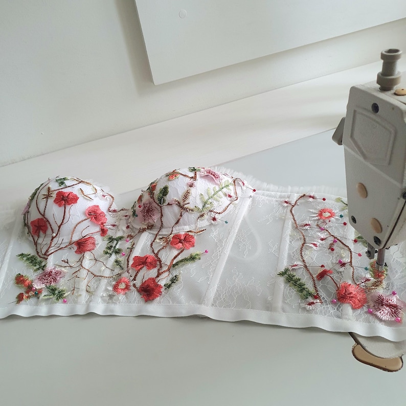 May include: White lace corset with floral embroidery in shades of pink, green, and red. The corset is being sewn on a sewing machine.
