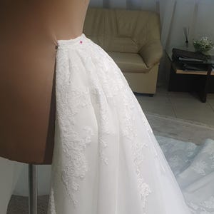 May include: A white wedding dress with intricate lace detailing is draped over a dress form. The dress is pinned in place with a pink pin, showcasing the delicate fabric and design. The dress is in the process of being tailored.