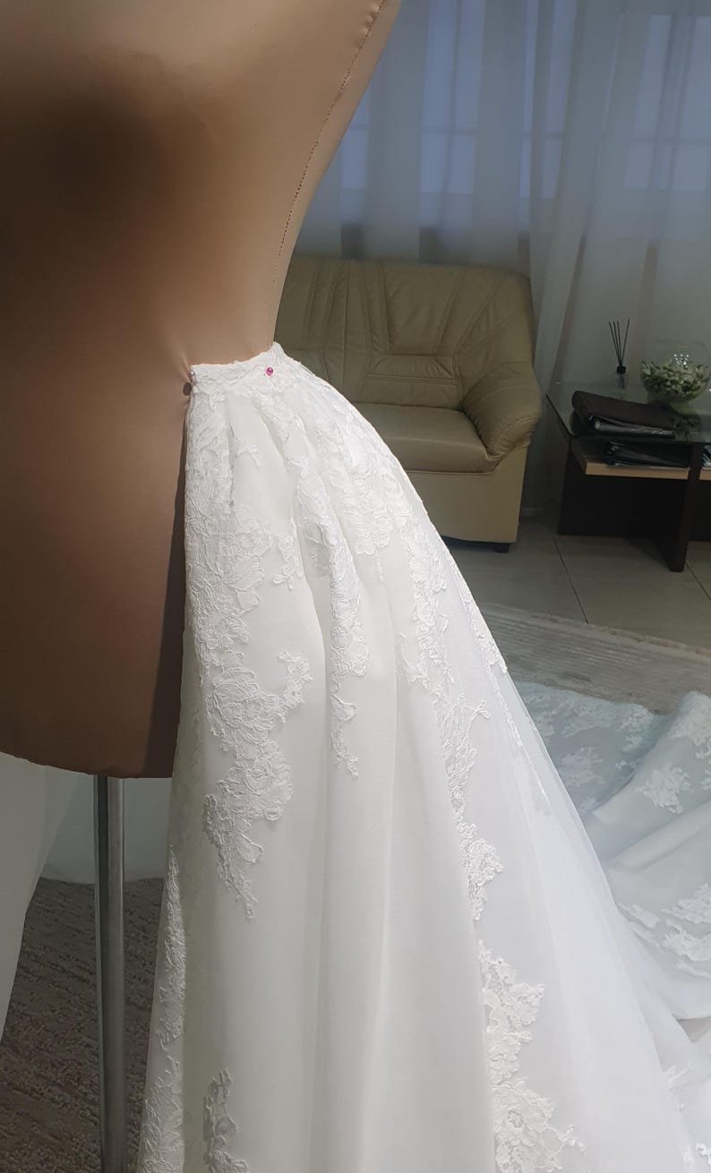 May include: A white wedding dress with intricate lace detailing is draped over a dress form. The dress is pinned in place with a pink pin, showcasing the delicate fabric and design. The dress is in the process of being tailored.