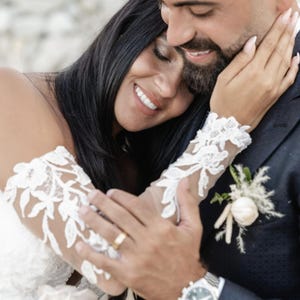 May include: A couple embraces, smiling on their wedding day. The bride wears a white lace sleeve dress, and the groom wears a navy suit with a boutonniere. A gold wedding band is visible. The image captures a moment of joy and affection.