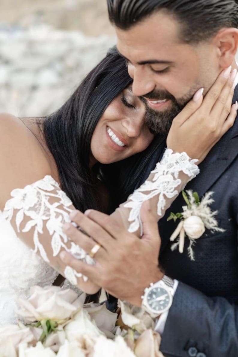 May include: A couple embraces, smiling on their wedding day. The bride wears a white lace sleeve dress, and the groom wears a navy suit with a boutonniere. A gold wedding band is visible. The image captures a moment of joy and affection.