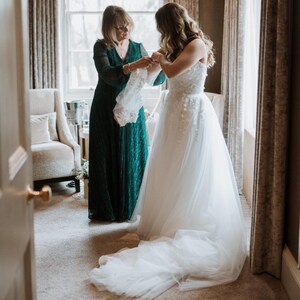 May include: A bride in a white strapless wedding gown with floral embroidery and a long train. A woman in a dark green dress assists the bride. The scene takes place in a room with a window and curtains.