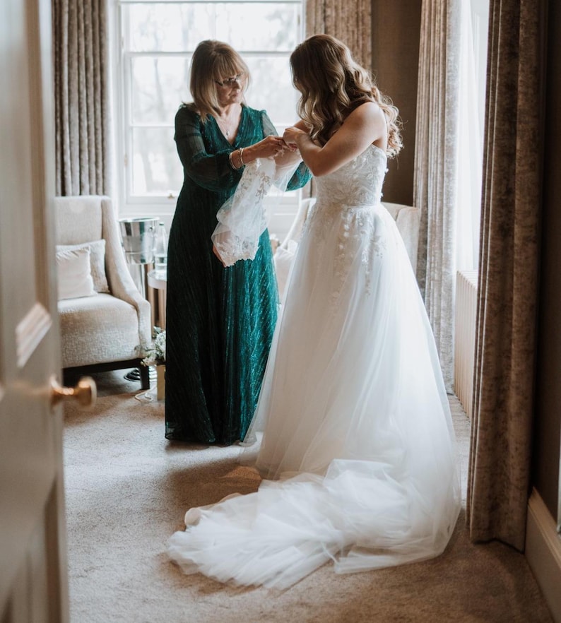 May include: A bride in a white strapless wedding gown with floral embroidery and a long train. A woman in a dark green dress assists the bride. The scene takes place in a room with a window and curtains.