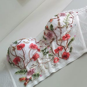May include: White lace corset with floral embroidery in shades of red, pink, and green. The corset has a boned structure and a satin trim.