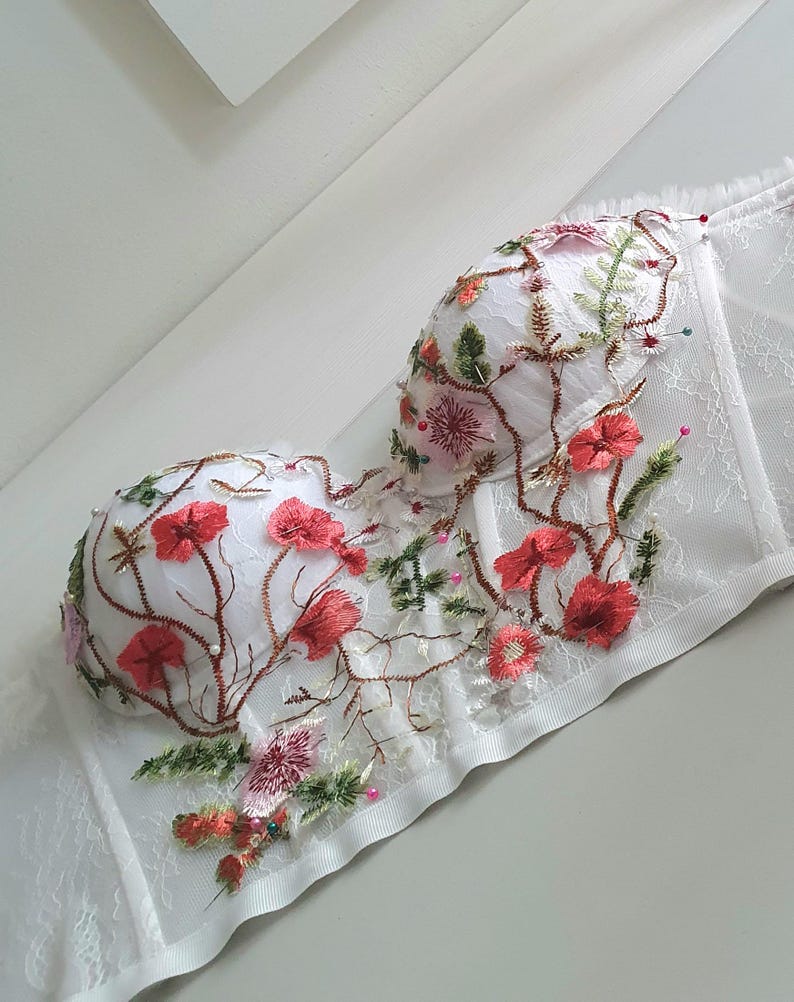 May include: White lace corset with floral embroidery in shades of red, pink, and green. The corset has a boned structure and a satin trim.
