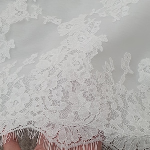 May include: Close-up of white lace fabric with floral patterns. The delicate lace features intricate designs and a scalloped edge. The fabric appears to be semi-sheer, with a soft, textured appearance. This textile is suitable for bridal wear or other special occasion garments.