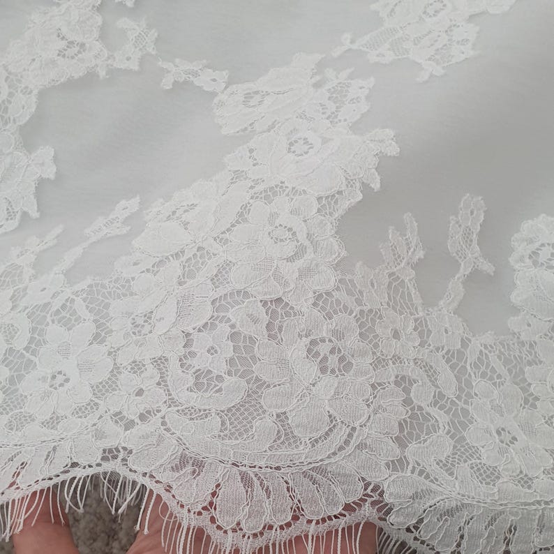 May include: Close-up of white lace fabric with floral patterns. The delicate lace features intricate designs and a scalloped edge. The fabric appears to be semi-sheer, with a soft, textured appearance. This textile is suitable for bridal wear or other special occasion garments.
