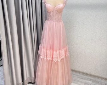 May include: A light pink formal gown on a mannequin. The bodice features a lace-up design with a sweetheart neckline and ruffled straps. The skirt is layered tulle with a lace trim detail. The dress is floor-length.