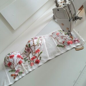 May include: White lace corset with floral embroidery in shades of pink, red, and green. The corset is being sewn on a white sewing machine.
