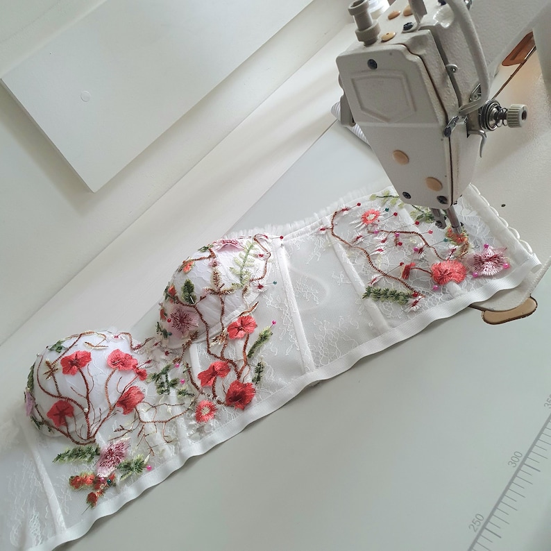 May include: White lace corset with floral embroidery in shades of pink, red, and green. The corset is being sewn on a white sewing machine.