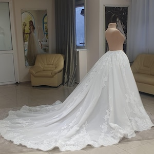 May include: A white wedding dress with a long train, displayed on a mannequin. The dress features intricate lace detailing along the hem and train edges. The dress is in a bridal shop setting, with a mirror and seating visible in the background.