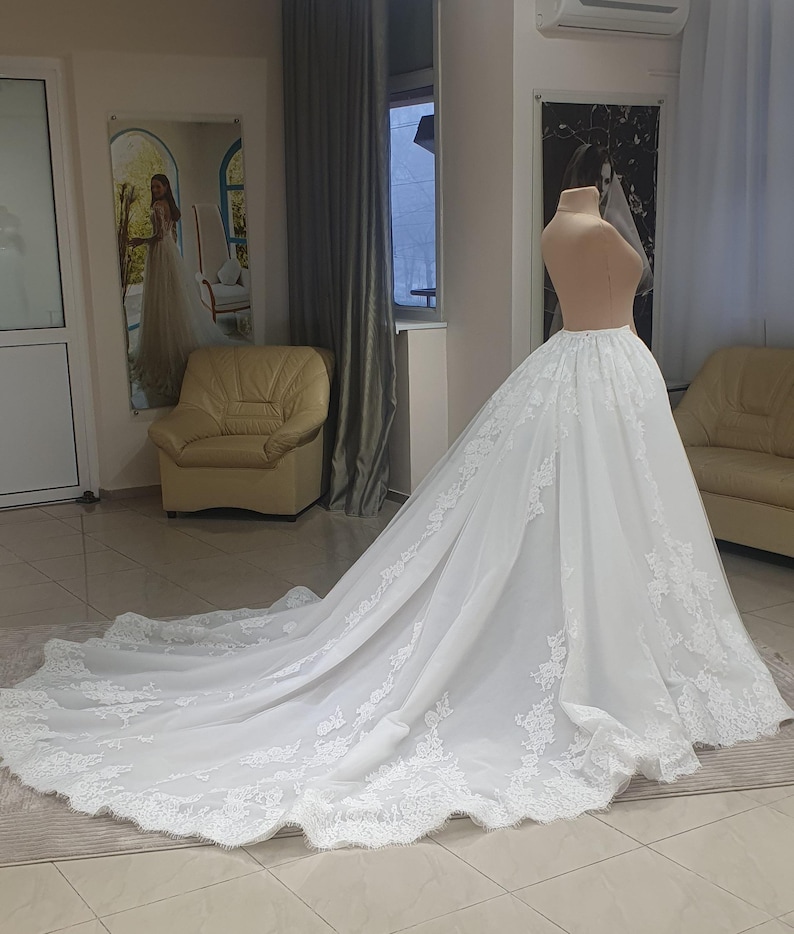 May include: A white wedding dress with a long train, displayed on a mannequin. The dress features intricate lace detailing along the hem and train edges. The dress is in a bridal shop setting, with a mirror and seating visible in the background.
