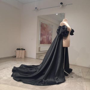 May include: A black satin gown with a dramatic train and puffed sleeves is displayed on a mannequin. The dress is floor-length and has a flowing, elegant design. The mannequin is in a well-lit room with a neutral background.