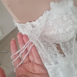 May include: A white wedding dress with a lace bodice and a beaded strap. The dress has a fitted bodice and a flowing skirt.