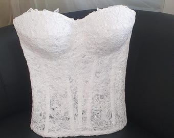 May include: A white strapless corset top featuring intricate floral lace embroidery and subtle sequin embellishments. The garment has a sweetheart neckline, structured boning throughout the bodice, and a scalloped hemline along the top edge.