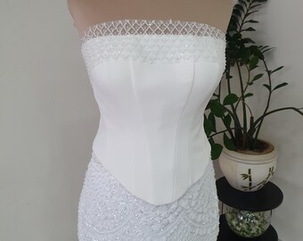 May include: A strapless white corset top featuring a structured bodice with vertical seams and a sheer upper panel adorned with intricate white beadwork and sequins. The garment transitions into a heavily beaded white skirt with scalloped patterns.