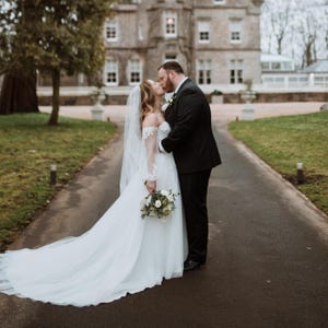May include: A couple shares a kiss on a paved path, celebrating their wedding. The bride wears a white, off-the-shoulder gown with a long train and veil, holding a bouquet. The groom is in a black suit. A large stone building is in the background.