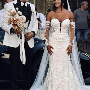 May include: A bride in a white, floral lace wedding dress with long, sheer sleeves and a sweetheart neckline. She is standing next to a man in a white tuxedo jacket, black pants, and a bow tie, holding a bouquet of flowers.
