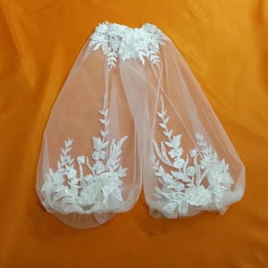 May include: A white bridal veil with floral embroidery. The veil is made of sheer fabric and features detailed white floral and leaf patterns along the edges and top. The veil is displayed against an orange background.