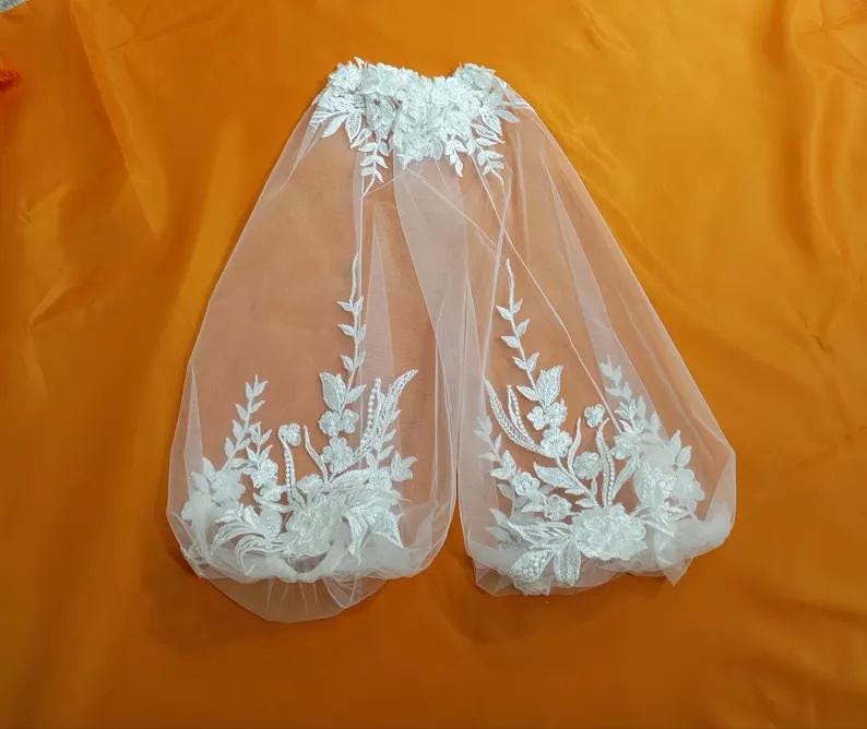 May include: A white bridal veil with floral embroidery. The veil is made of sheer fabric and features detailed white floral and leaf patterns along the edges and top. The veil is displayed against an orange background.