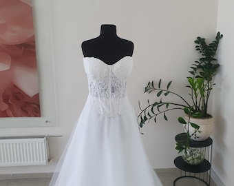May include: A white strapless gown featuring a structured corset-style bodice with intricate lace detailing and a sweetheart neckline. The bodice transitions into a full-length, layered tulle skirt that flows downward from the waistline.