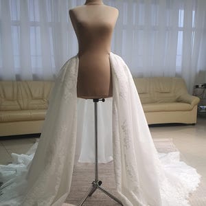 May include: A white bridal skirt with intricate lace detailing is draped over a mannequin. The skirt is displayed on a metal stand, showcasing its flowing design. The background features a neutral-toned room with a sofa and curtains.