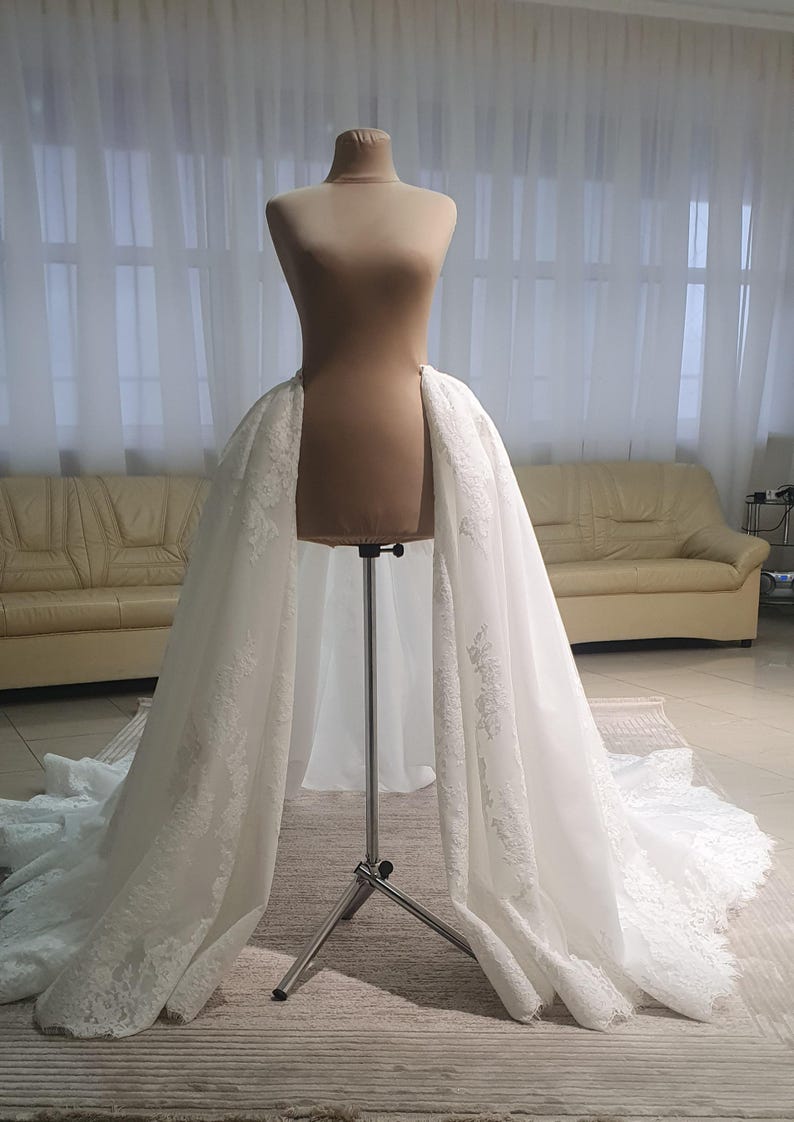 May include: A white bridal skirt with intricate lace detailing is draped over a mannequin. The skirt is displayed on a metal stand, showcasing its flowing design. The background features a neutral-toned room with a sofa and curtains.