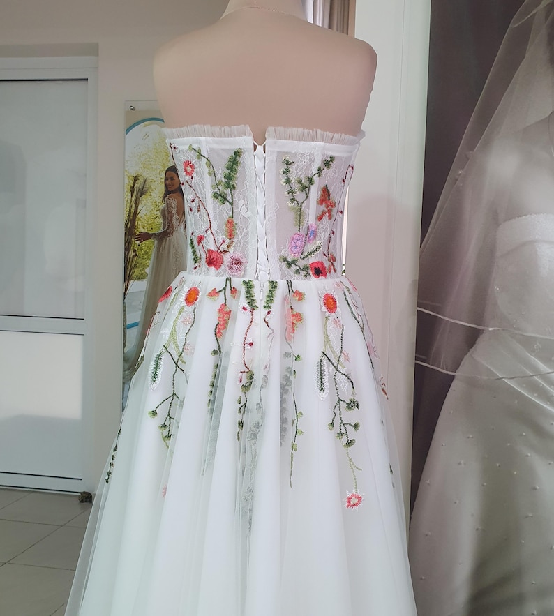 May include: A white strapless wedding dress with a tulle skirt and floral embroidery. The dress has a fitted bodice with a lace-up back.
