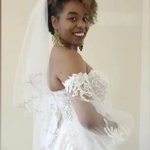 May include: A white wedding dress with a sweetheart neckline and lace detailing. The dress features off-the-shoulder sleeves and a tulle veil. The bride is wearing green earrings.