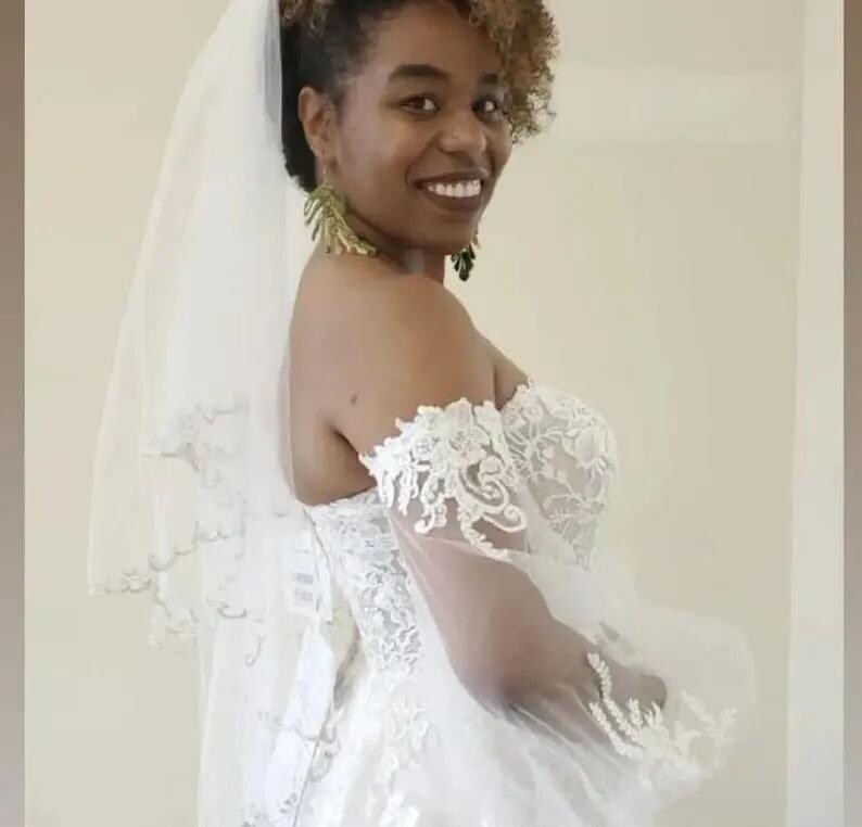 May include: A white wedding dress with a sweetheart neckline and lace detailing. The dress features off-the-shoulder sleeves and a tulle veil. The bride is wearing green earrings.