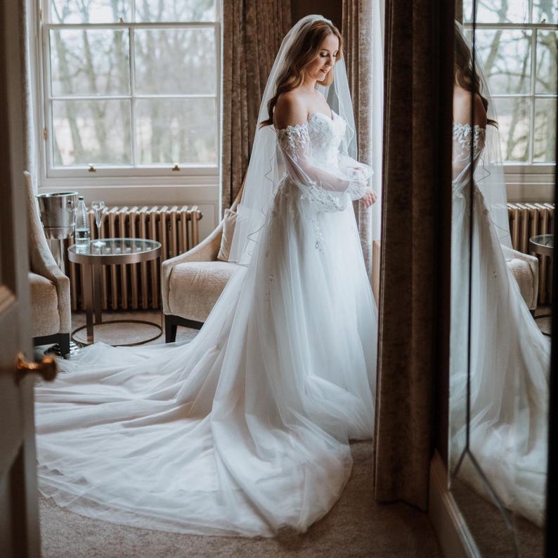 May include: A white off-the-shoulder wedding dress with a long train. The dress features delicate lace detailing on the bodice and sleeves. A sheer veil cascades down the back. The bride is standing in front of a mirror.