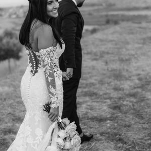 May include: A black and white photo of a couple walking outdoors. The woman wears a white, off-the-shoulder wedding dress with lace sleeves and holds a bouquet. The man is in a black suit. They are holding hands.