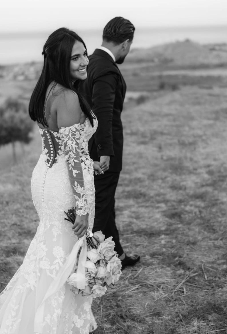 May include: A black and white photo of a couple walking outdoors. The woman wears a white, off-the-shoulder wedding dress with lace sleeves and holds a bouquet. The man is in a black suit. They are holding hands.