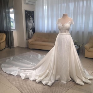 Op de afbeelding: Een witte trouwjurk op een mannequin. De jurk heeft een nauwsluitend lijfje met ingewikkelde kralen en een doorschijnende halslijn. De rok loopt uit in een lange sleep. De jurk wordt getoond in een lichte kamer.
