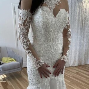 May include: A white wedding dress with intricate lace detailing and a mermaid silhouette. The dress features long, sheer sleeves with floral lace accents, a high neckline, and a bodice embellished with beads and embroidery. The dress is floor-length.