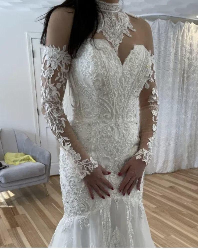 May include: A white wedding dress with intricate lace detailing and a mermaid silhouette. The dress features long, sheer sleeves with floral lace accents, a high neckline, and a bodice embellished with beads and embroidery. The dress is floor-length.