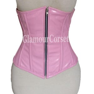 May include: A light pink corset with a front zipper closure. The corset has a glossy finish and vertical seams for shaping. The text "Glamour Corset" is printed on the front. The corset is displayed on a mannequin.